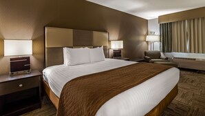 Quarto de hotel com uma cama grande, duas mesas de cabeceira com abajures, uma cadeira, uma escrivaninha com um computador e uma janela com cortinas.
