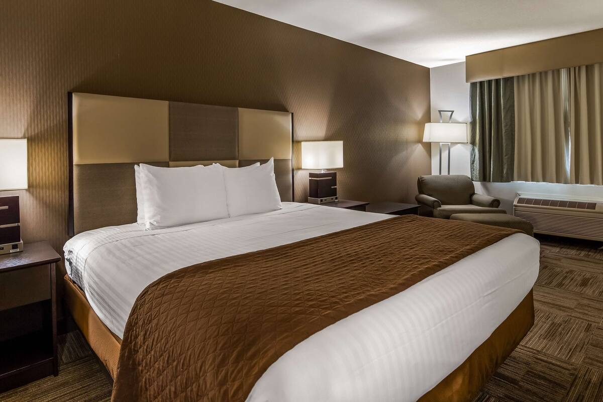 Ein Hotelzimmer mit einem großen Bett, zwei Nachttischen mit Lampen, einem Sessel, einem Schreibtisch mit Computer und einem Fenster mit Vorhängen.