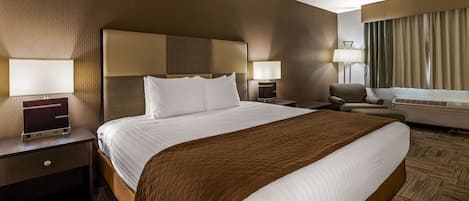 A hotel room with a large bed, two bedside tables with lamps, a chair, a desk with a computer, and a window with curtains.