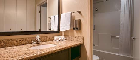 Un baño con lavamanos de encimera de granito, espejo, dispensador de toallas y ducha con cortina.