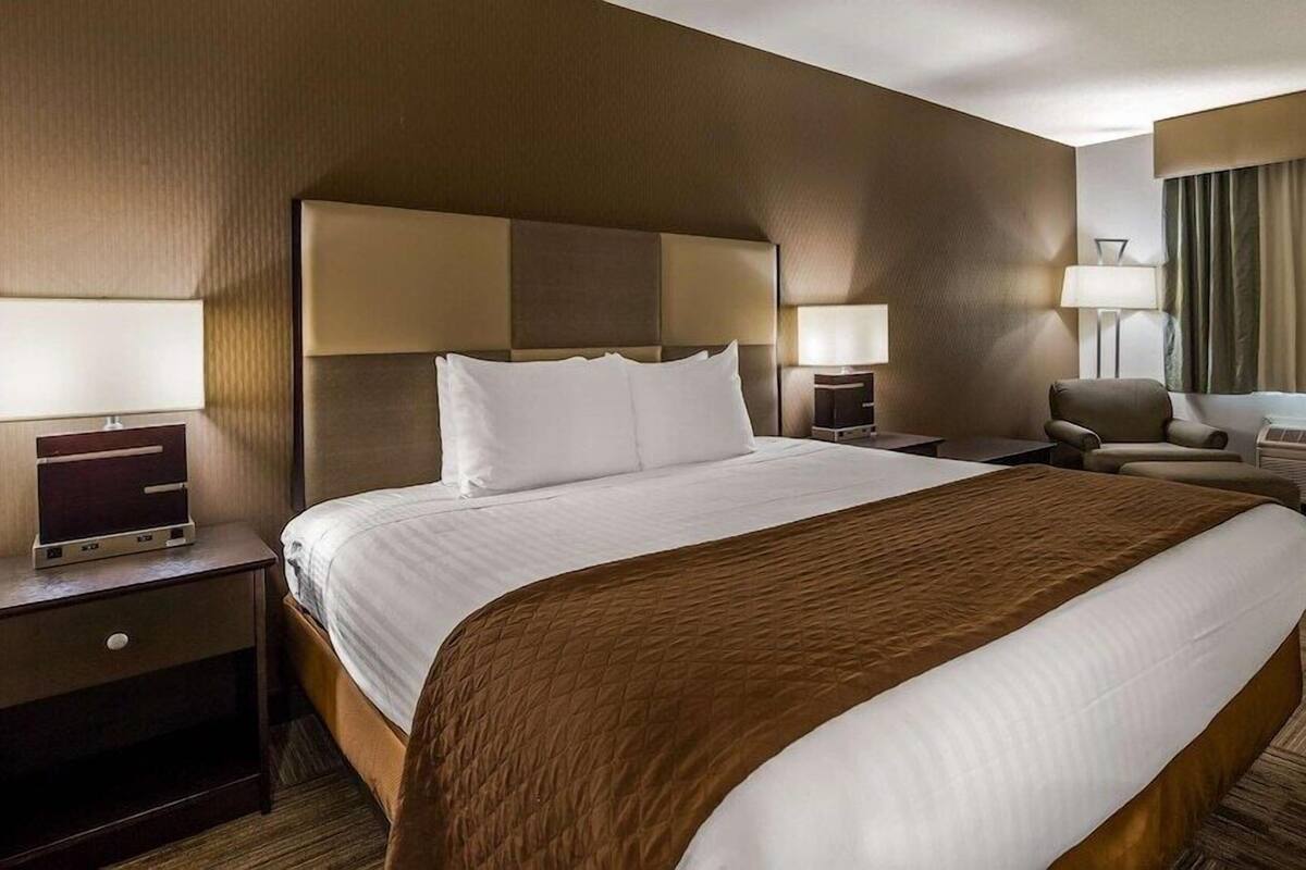 Ein Hotelzimmer mit einem großen Bett, zwei Nachttischen mit Lampen, einem Sessel, einem Schreibtisch und einem Fenster mit Vorhängen.