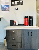 Mostrador de habitaciones de hotel con cafetera, una cafetera roja Folgers y una cafetera negra. Hay un cartel en la pared y un cesto de basura en la esquina.