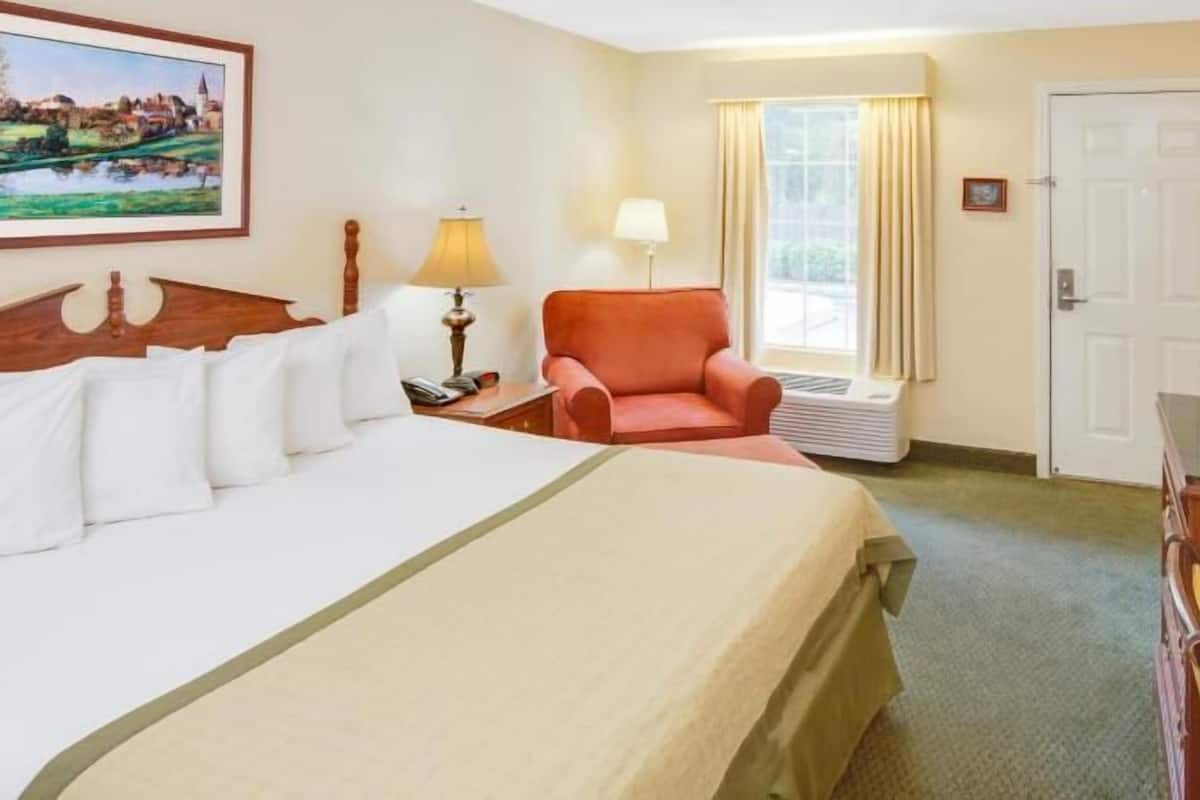Quarto de hotel com cama, poltrona, televisão, luminária, quadro e janela.