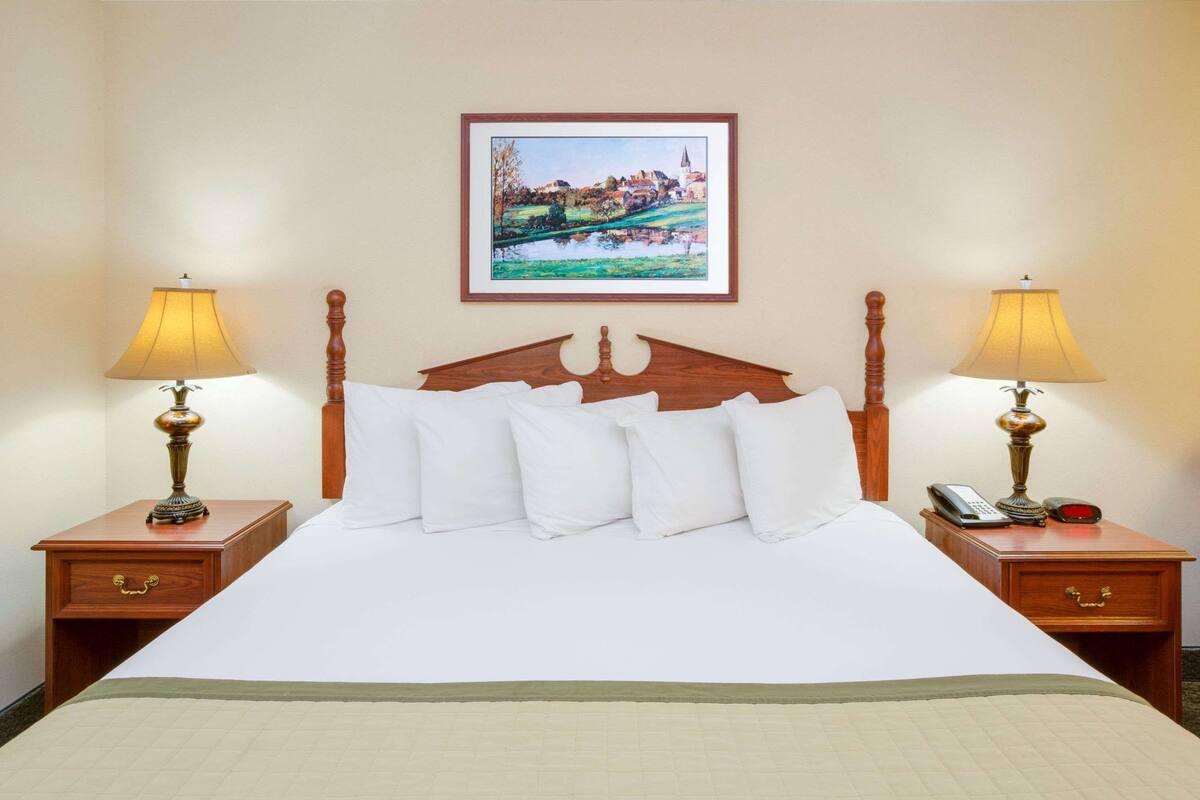Cama bem arrumada com lençóis brancos, duas mesas de cabeceira com abajures, um quadro na parede e uma cabeceira de madeira.