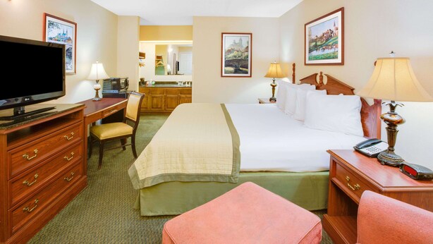 A hotel room with a bed, a desk with a television, a chair, a nightstand with a lamp, and a bathroom visible in the background. - Baymont by Wyndham Ozark (Ozark)