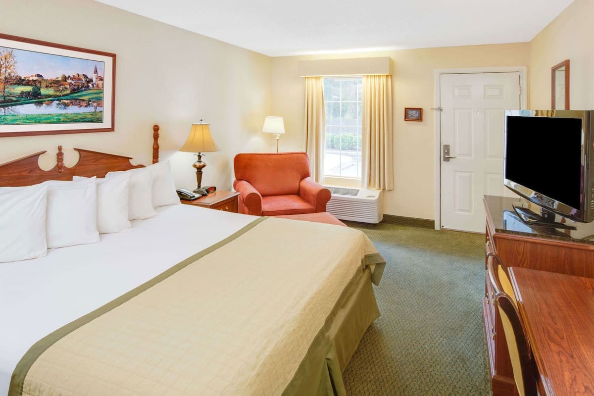 Quarto de hotel com cama, uma cadeira, televisão, janela com cortinas e um quadro na parede.