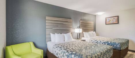 Habitación de hotel con dos camas, un sillón verde, una mesita de noche con lámpara y una obra de arte colgada en la pared.
