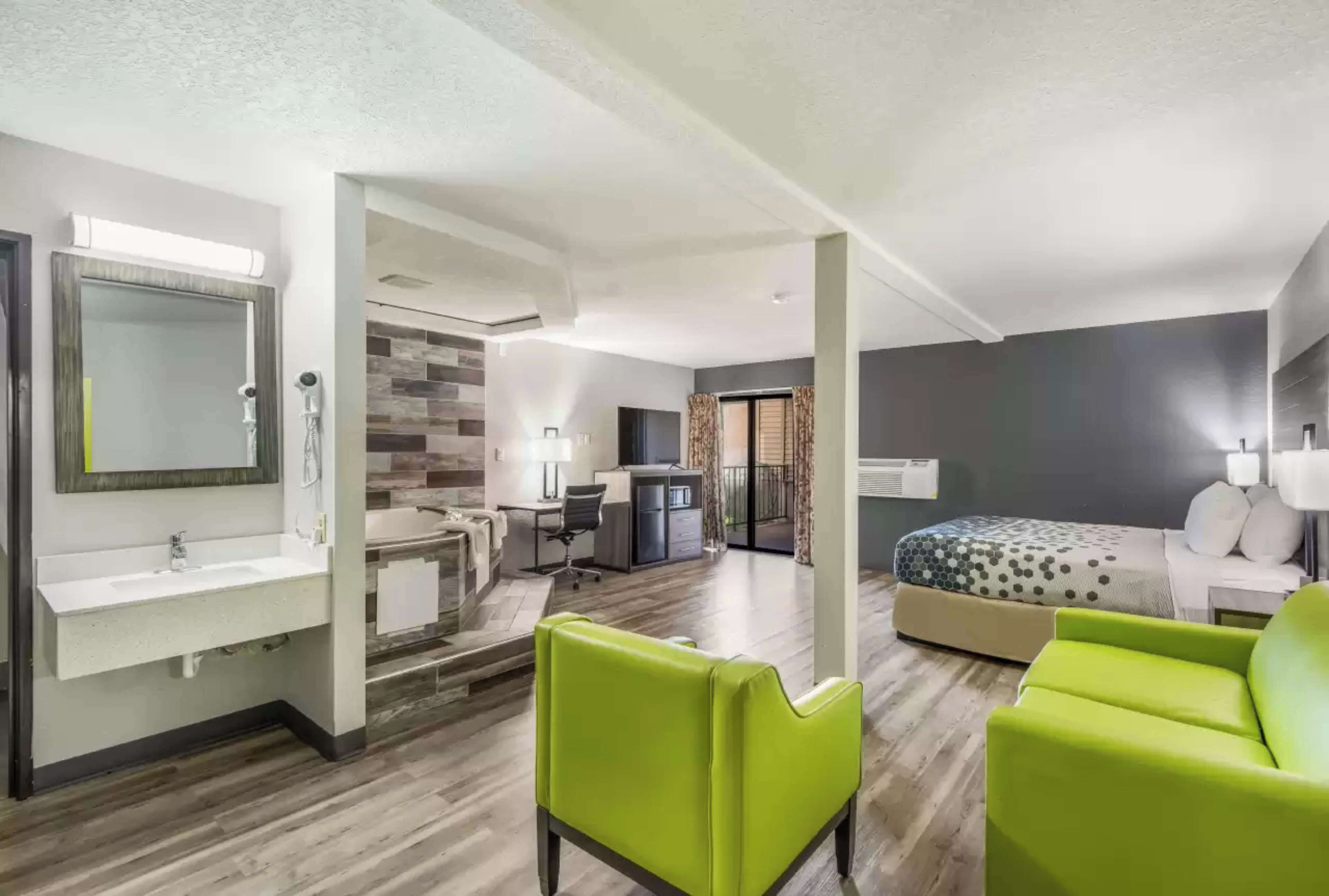 A modern hotel room with a large bed, a desk, a chair, and a bathroom with a sink and mirror.