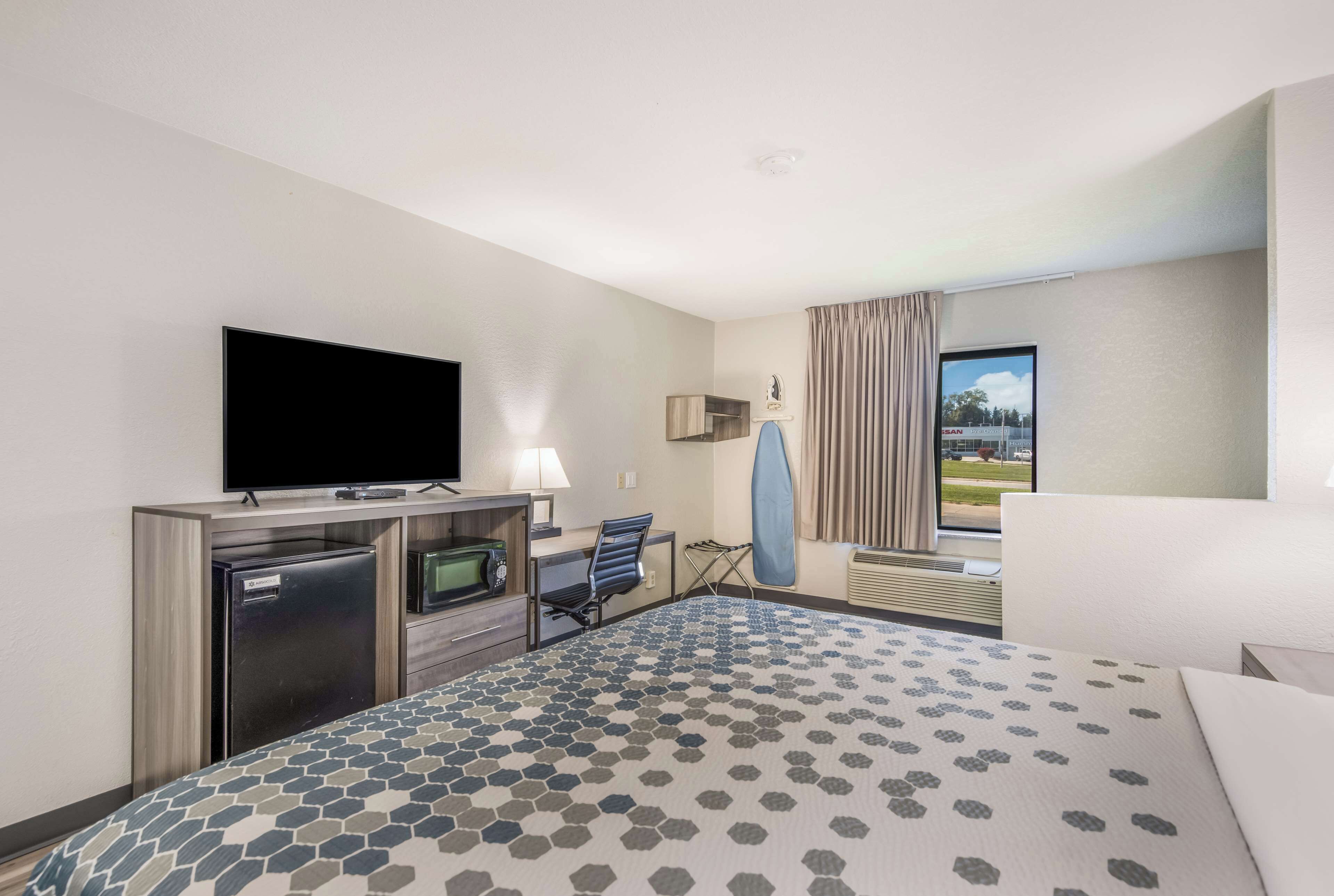 A hotel room with a bed, a desk, a television, a refrigerator, and a window with curtains.
