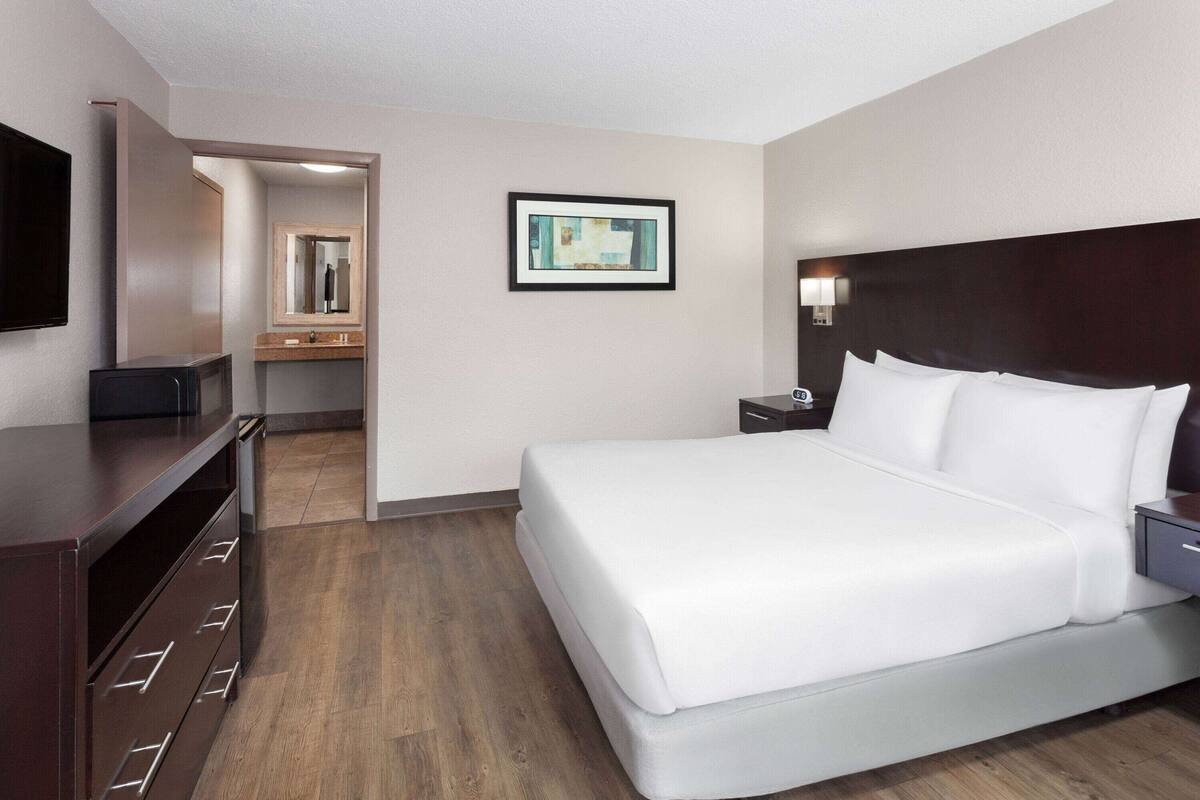 A hotel room with a large bed, a desk with drawers, a flat-screen TV, a framed picture on the wall, and a bathroom visible through an open door.