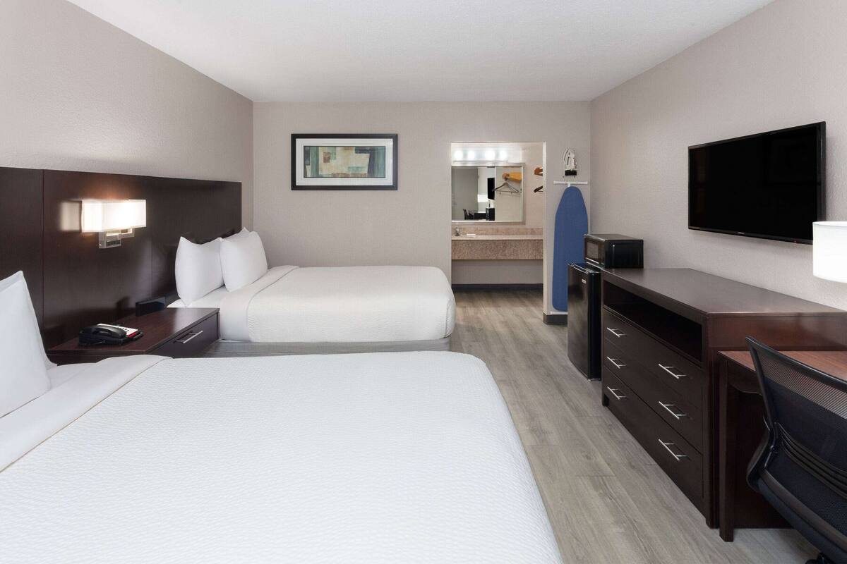 A hotel room with two beds, a desk, a dresser, a TV, a mirror, and a bathroom visible in the background.