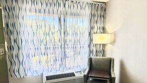 Quarto de hotel com cortina estampada, uma cadeira, mesa de cabeceira e luminária fixa na parede.