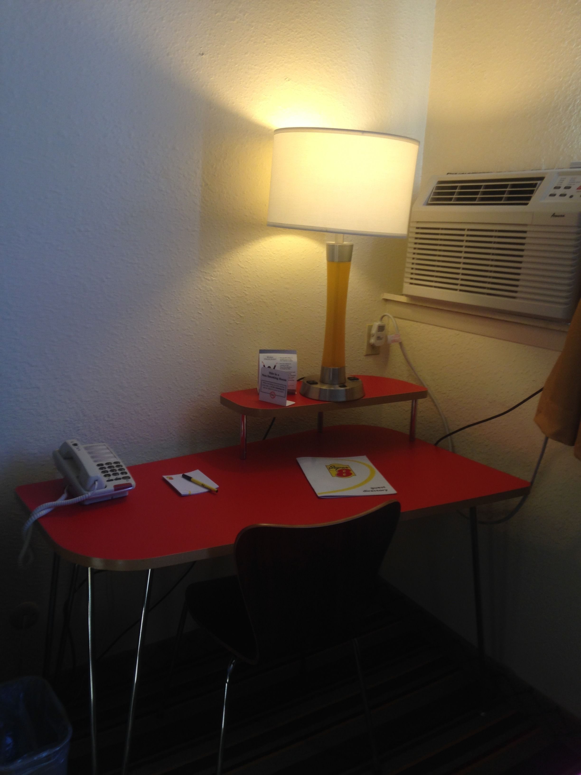 A small hotel room with a red desk, a lamp, a phone, and an air conditioner.