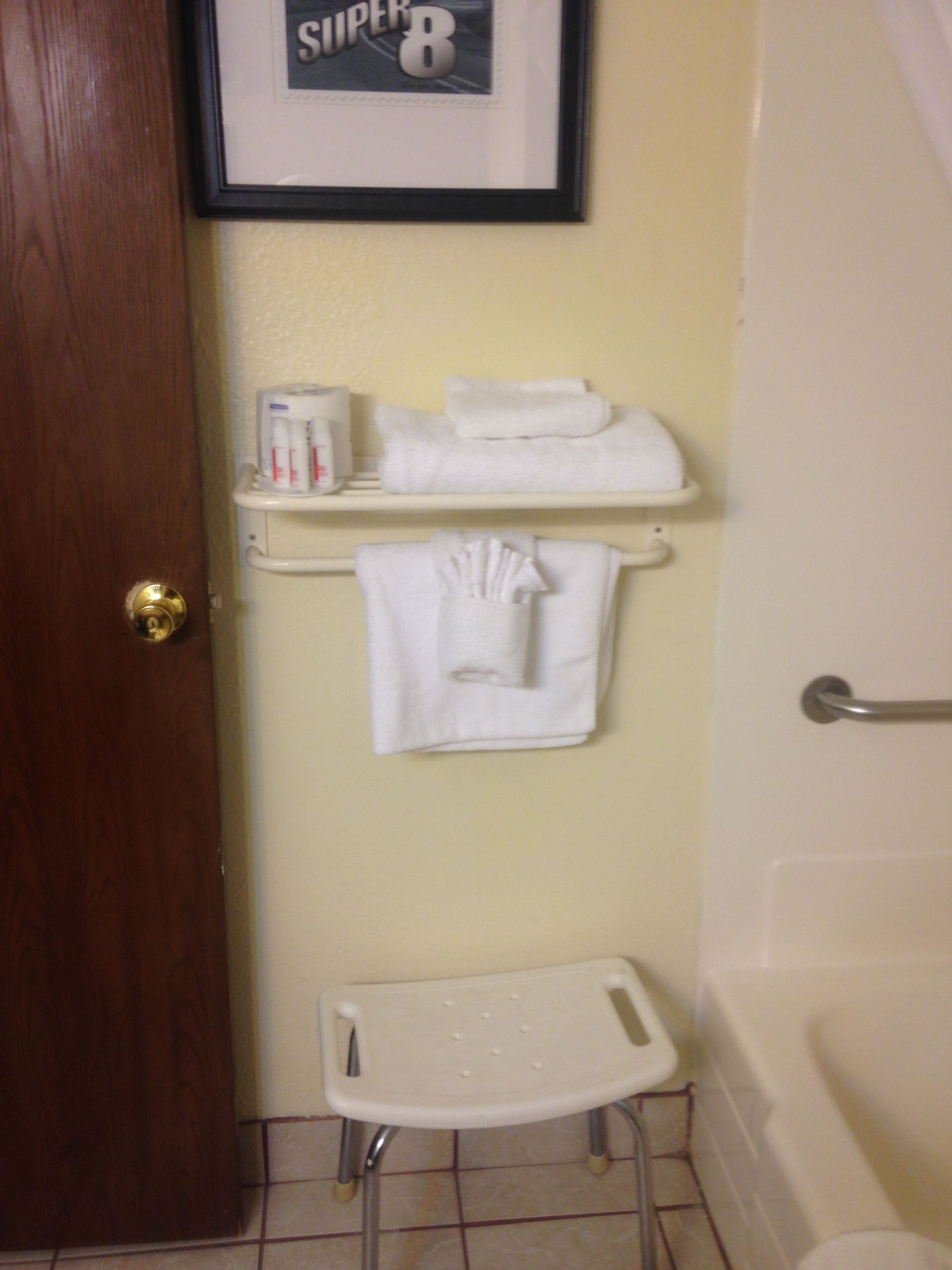 Une salle de bain avec une porte en bois, un tableau encadré au mur, une étagère avec des serviettes, un porte-rouleau de papier toilette blanc et un petit tabouret blanc.