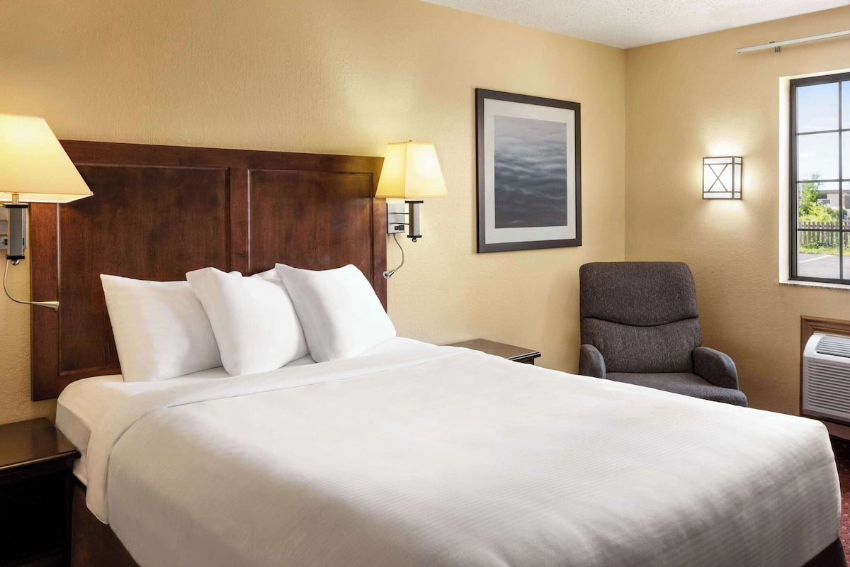 A hotel room with a large bed, two bedside lamps, a chair, a framed picture on the wall, and a window with a view.