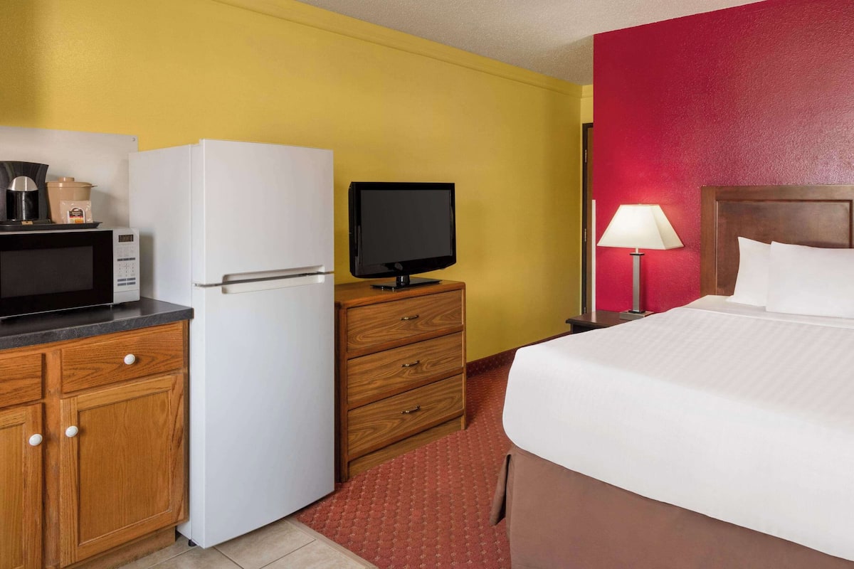 Une chambre d’hôtel avec un lit, un bureau doté d’un micro-ondes, un réfrigérateur, une télévision, une table de chevet avec une lampe et un mur rouge.