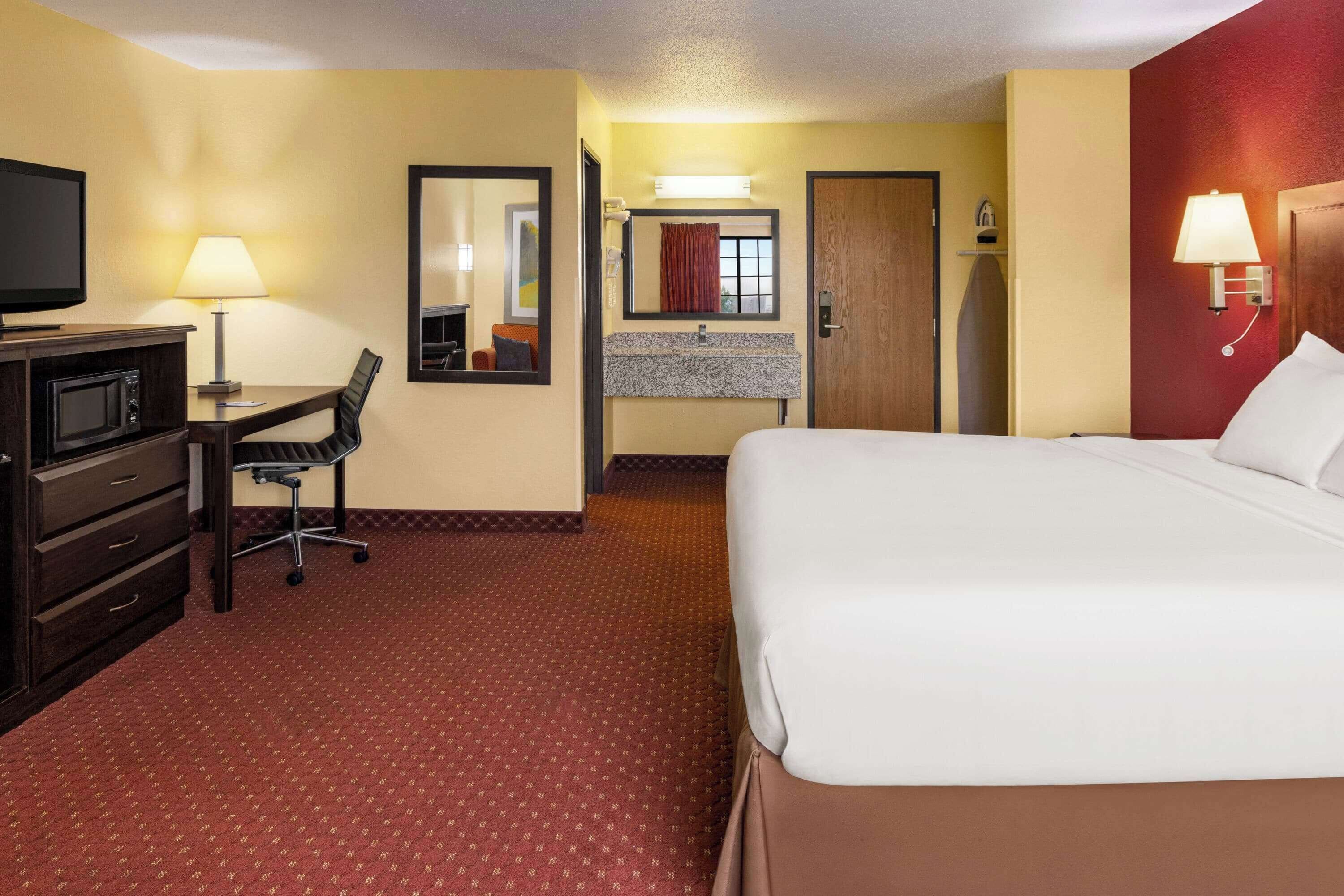 Habitación de hotel con cama, escritorio y silla, televisor, espejo, baño con lavamanos y ducha, y alfombra roja.