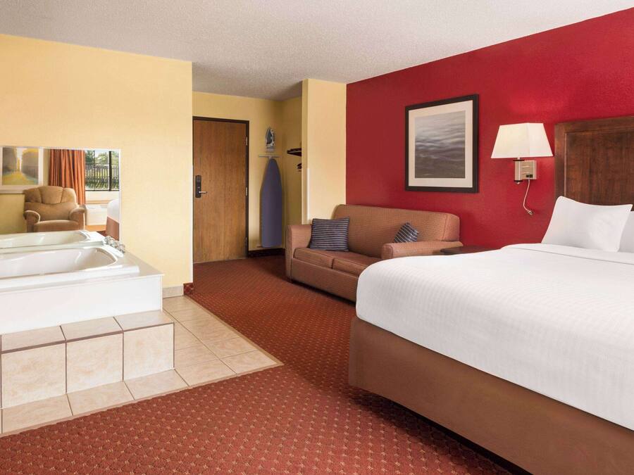 Quarto de hotel com uma cama grande, um sofá, uma poltrona, uma banheira e uma parede de destaque vermelha.