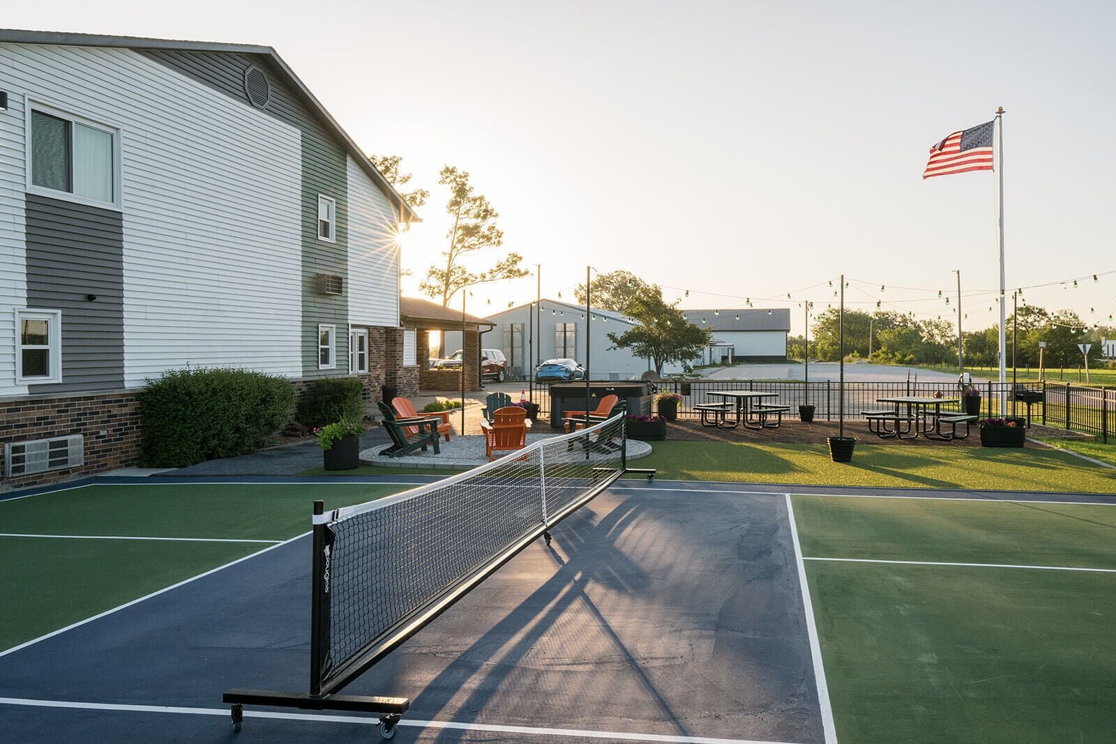 Un campo da tennis all'aperto con rete, area relax con sedie e un albero con bandiera americana.