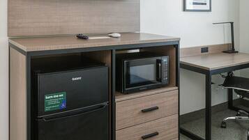 A compact hotel room with a flat-screen TV mounted on the wall, a minibar with a refrigerator and microwave, a desk with a chair, and a framed poster on the wall.