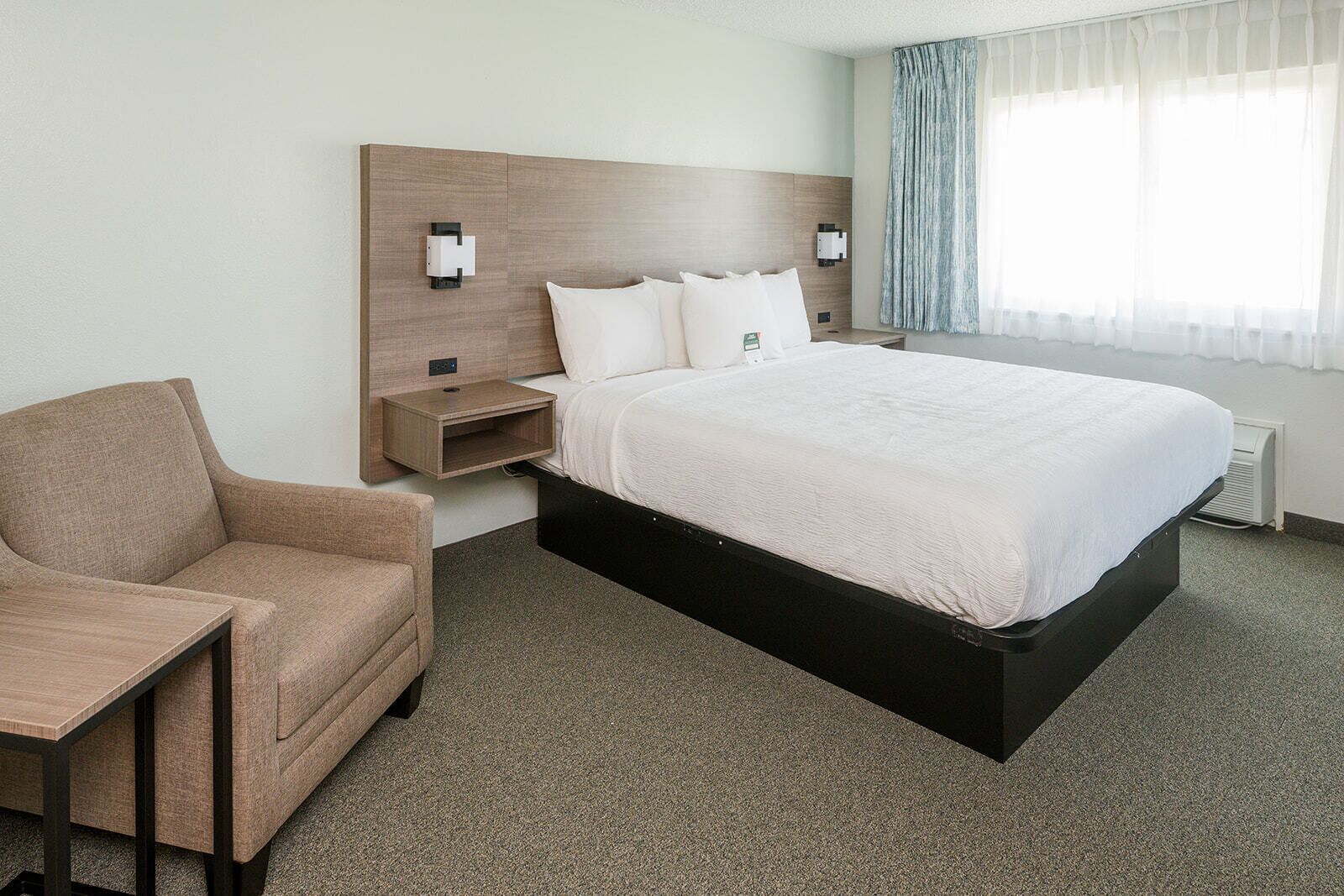 Une chambre d’hôtel avec un lit, une chaise, une table de chevet et une lampe fixée au mur.