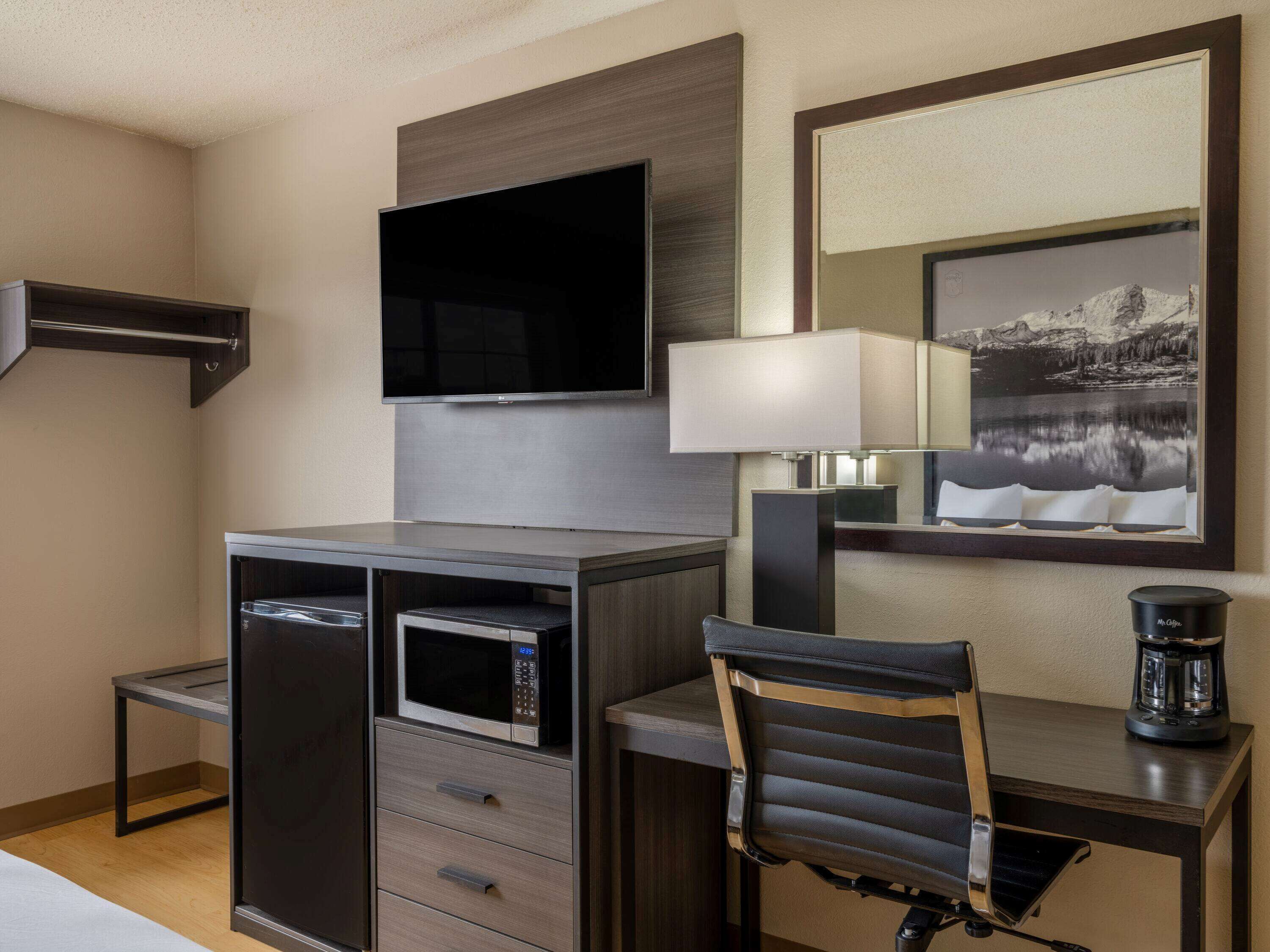 A hotel room with a flat-screen TV mounted on the wall, a desk with a lamp, a microwave, and a coffee maker.