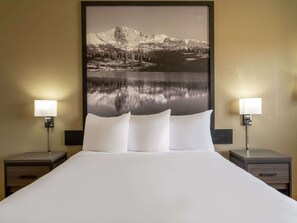 Une chambre d’hôtel avec un grand lit, deux tables de chevet avec des lampes et un grand tableau encadré représentant un paysage de montagne accroché au mur.
