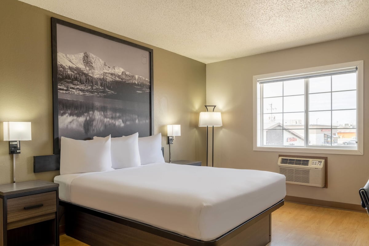 A hotel room with a large bed, two bedside tables with lamps, a large window, and a wall-mounted air conditioner.