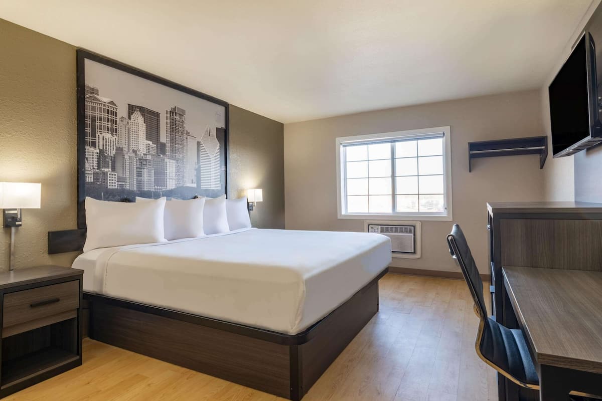 A hotel room with a large bed, a desk with a chair, a window, and a cityscape mural on the wall.