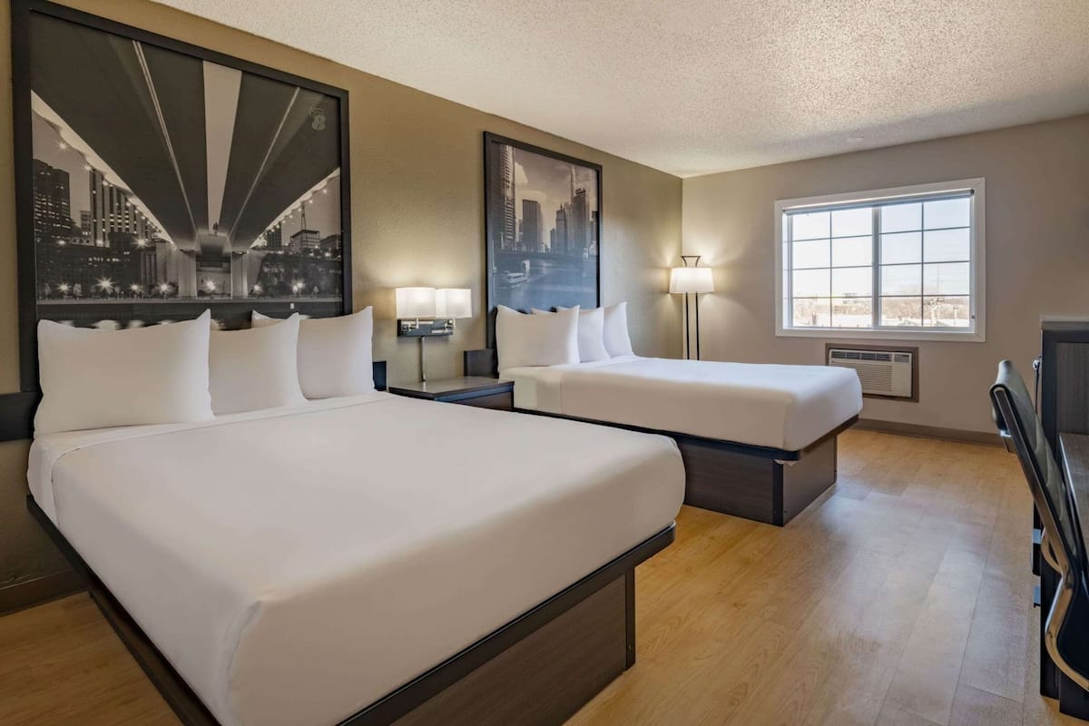 A hotel room with a large bed, a smaller bed, a desk, a chair, and a window with a city view.