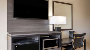 A compact hotel room with a flat-screen TV mounted on the wall, a desk with a lamp, a microwave, and a chair. - Super 8 by Wyndham Clinton (Clinton)