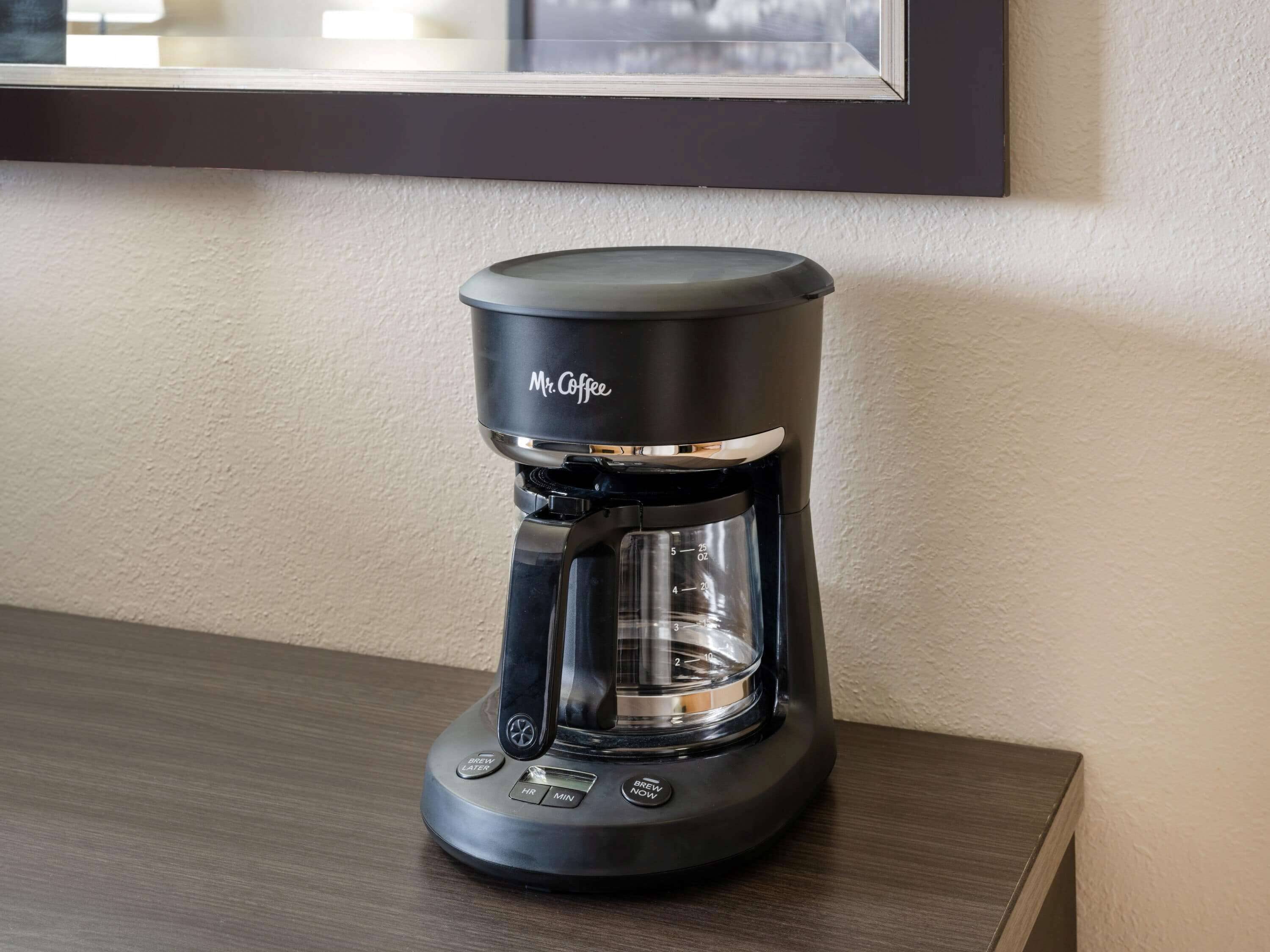 Una cafetera Mr. Coffee sobre una mesa de madera, frente a una pared con un cuadro enmarcado encima.