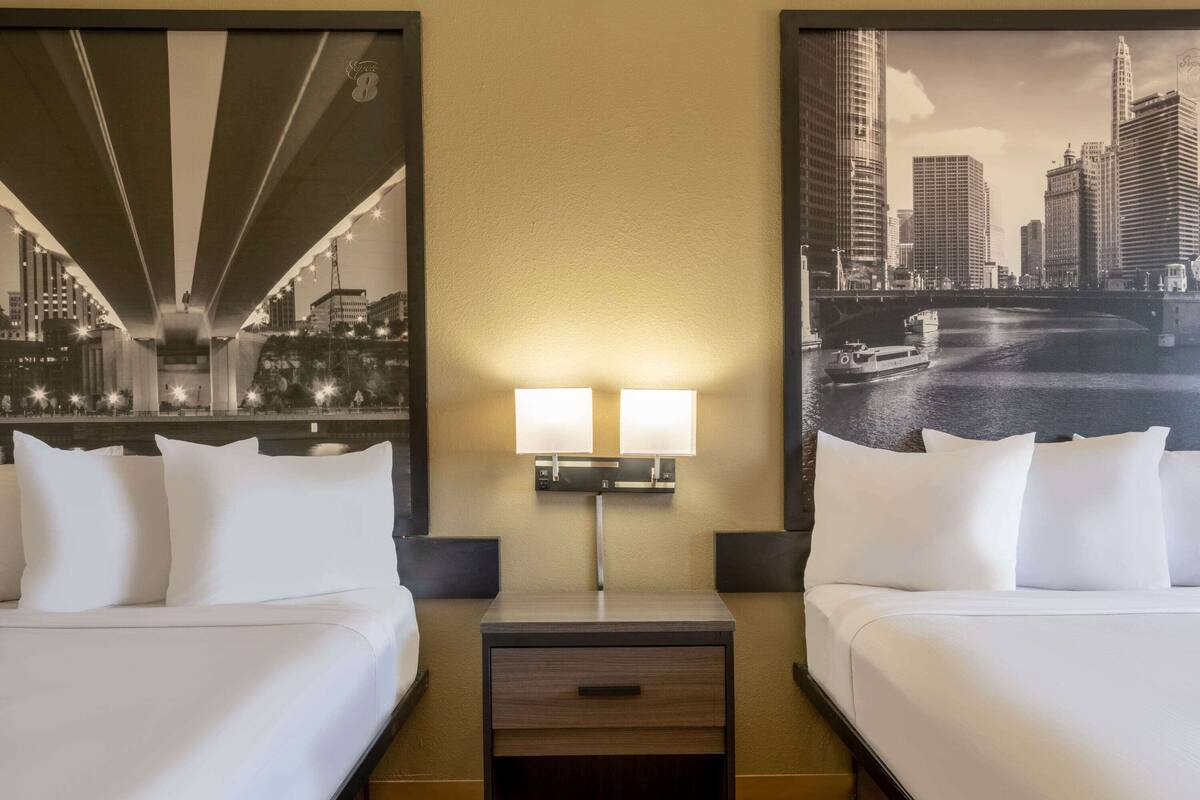 A hotel room with two beds, a nightstand with a lamp, and two large cityscape posters on the walls.