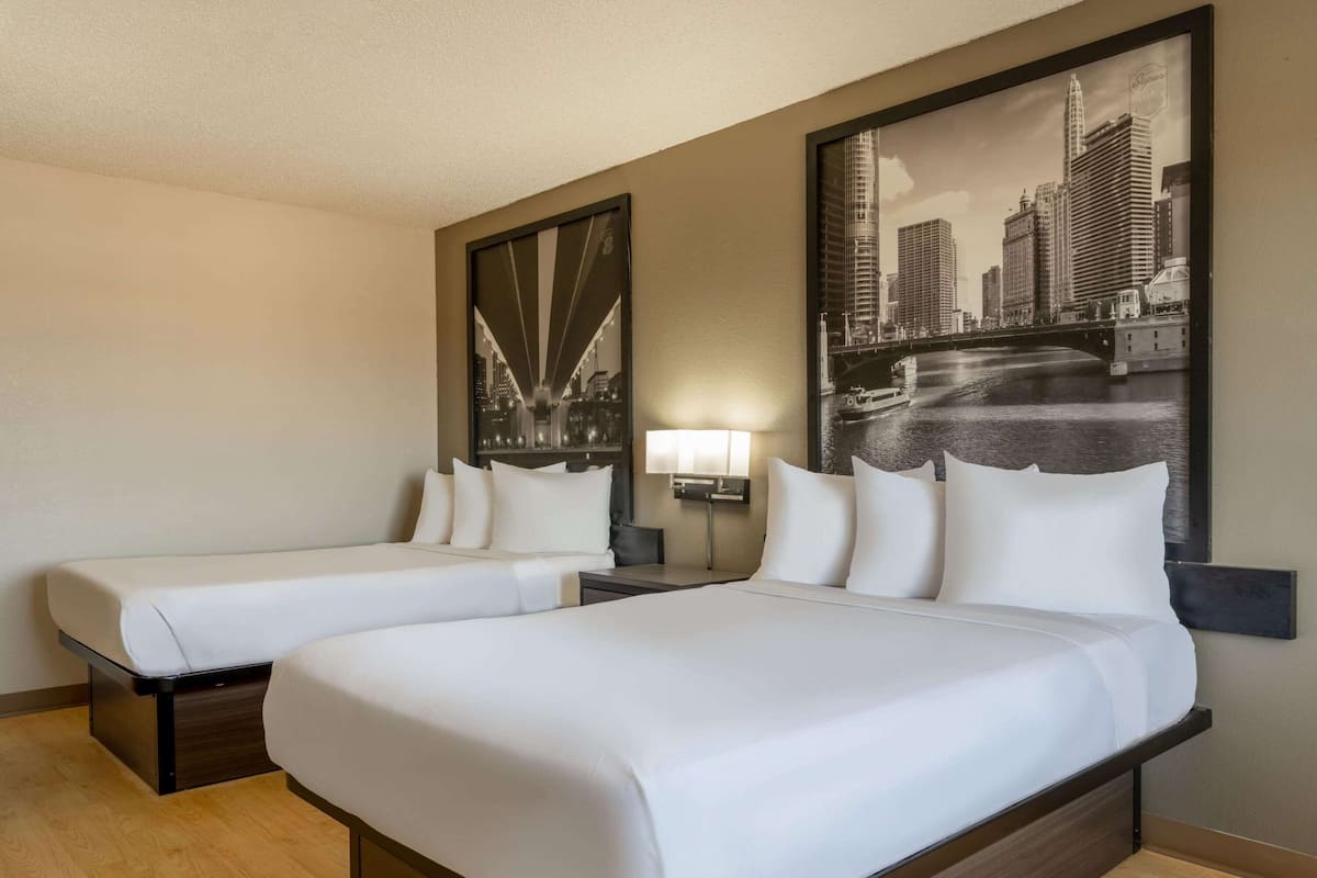 A hotel room with two beds, a large cityscape painting on the wall, a nightstand with a lamp, and a wooden floor.