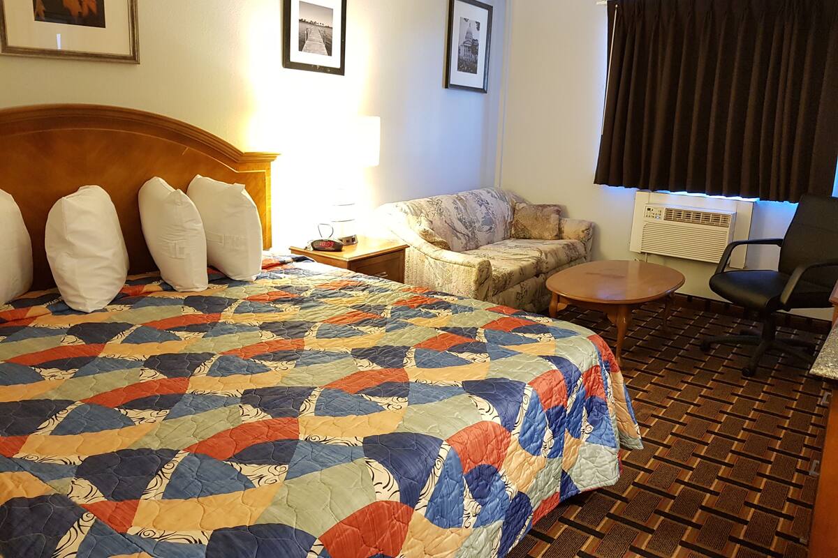 A hotel room with a large bed, a sofa, a small table, and a chair. There are framed pictures on the wall and an air conditioning unit.