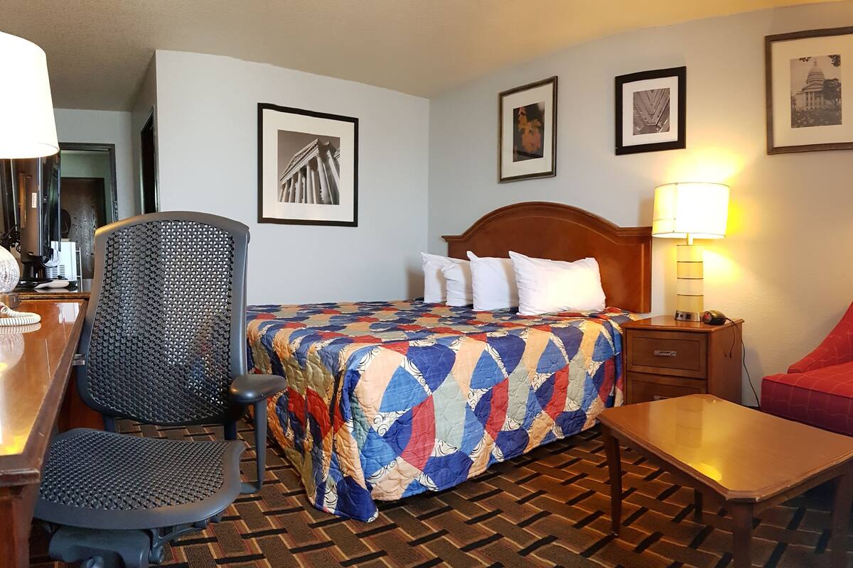 A hotel room with a bed, a desk with a chair, a red armchair, and a table. There are framed pictures on the wall and a lamp on the nightstand.