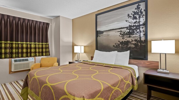 Een hotelkamer met een groot bed, een raam met gordijnen, een wandairconditioner, een bedlamp en een schilderij van een landschap.