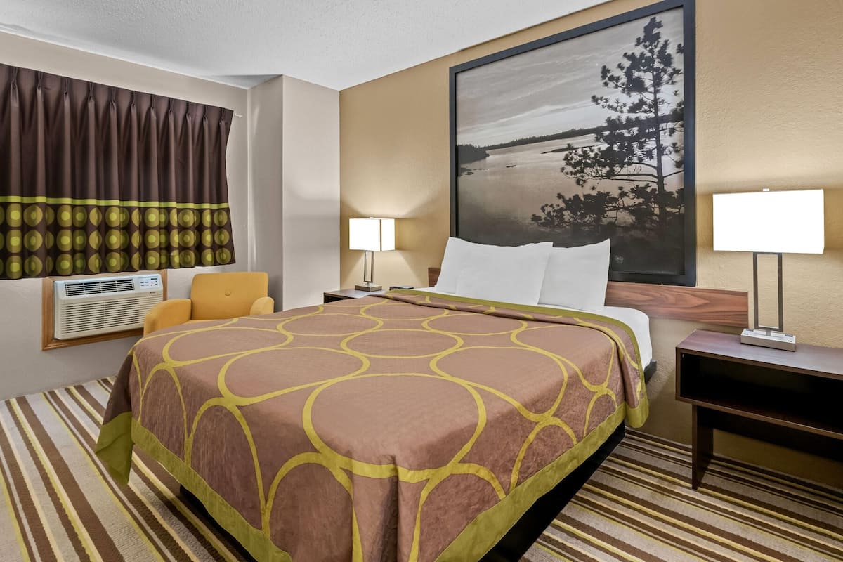 Une chambre d’hôtel avec un grand lit, une fenêtre avec des rideaux, un climatiseur fixé au mur, une lampe de chevet et un tableau représentant un paysage.