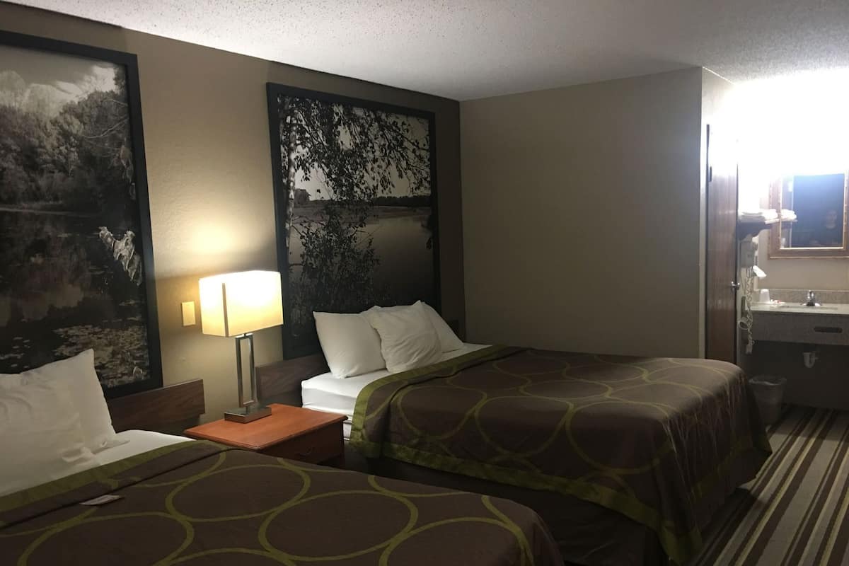 Une chambre d’hôtel avec deux lits, une table de chevet avec une lampe, une commode avec un miroir et une salle de bain visible à travers une porte ouverte.