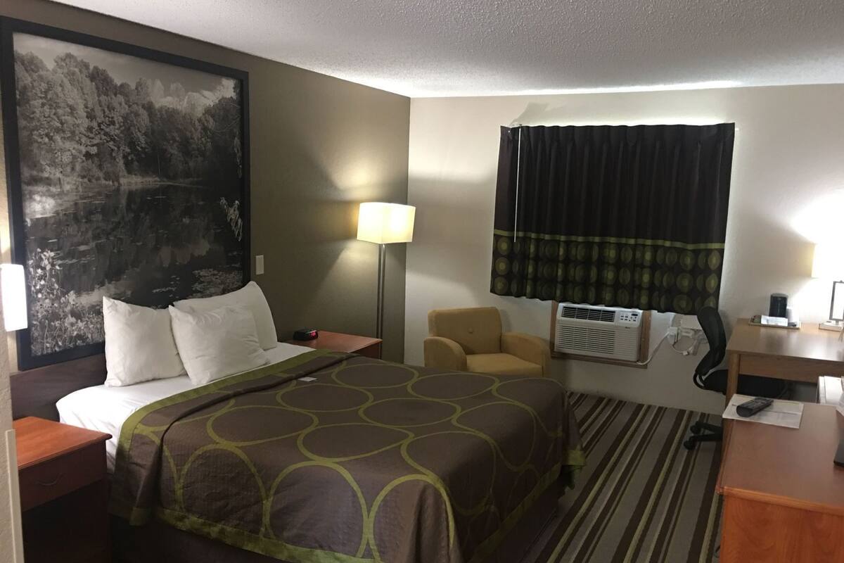 Une chambre d’hôtel avec un lit, une chaise, un bureau, une lampe, un climatiseur et un grand tableau au mur.