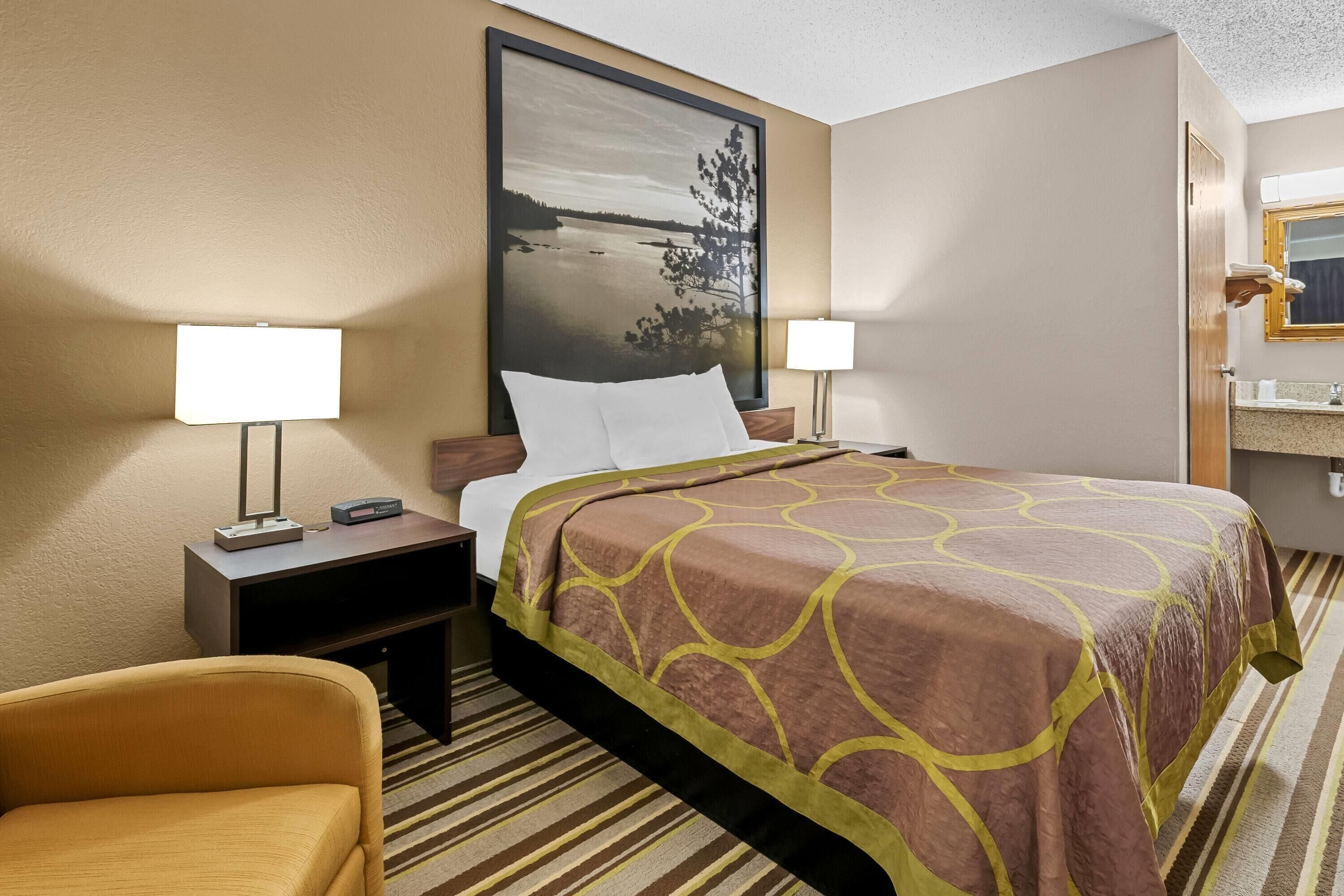 A hotel room with a large bed, two bedside tables with lamps, a chair, a large framed picture on the wall, and a bathroom visible through an open door.
