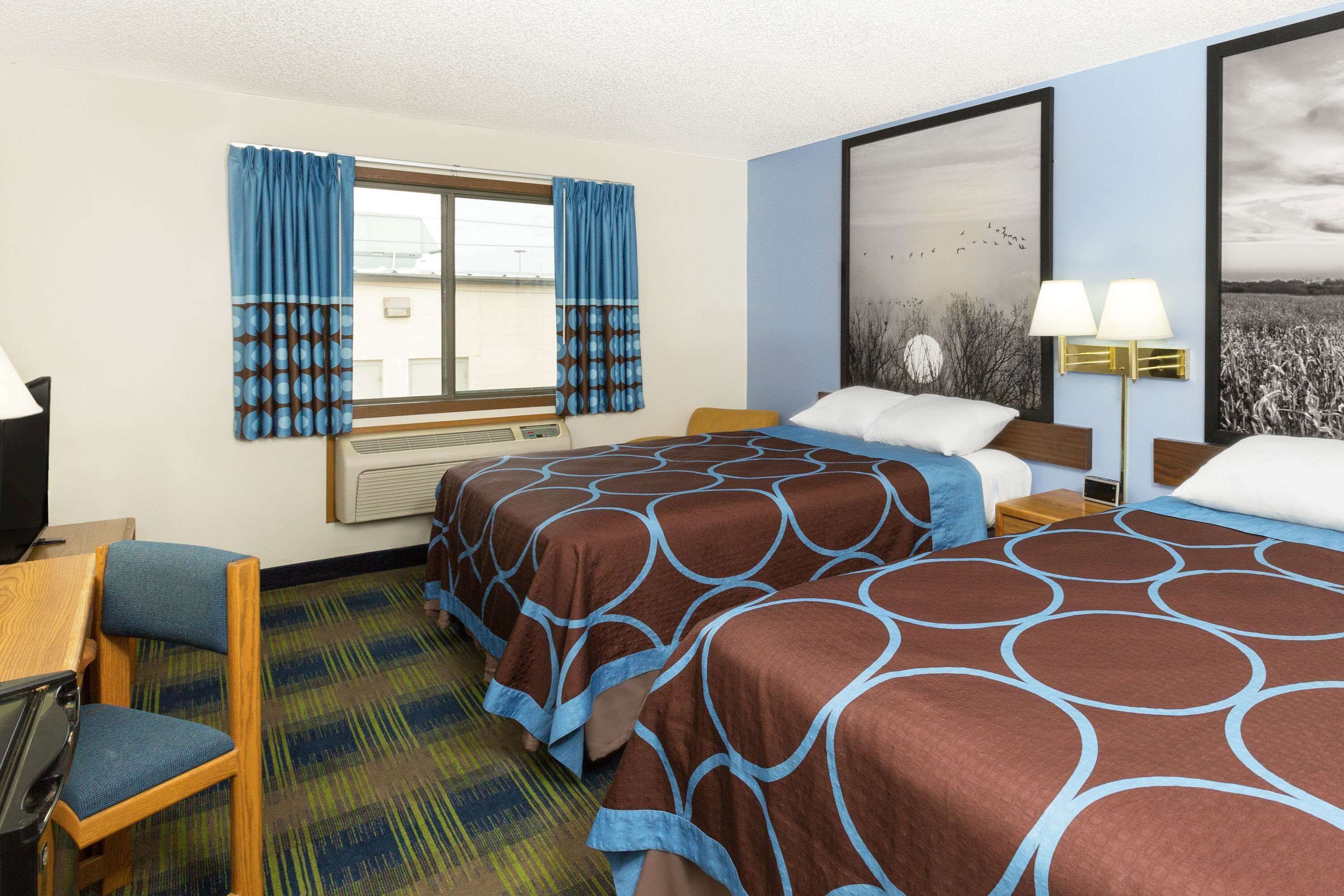 A hotel room with two beds, a desk, a chair, a window with curtains, a lamp, and a large painting on the wall.