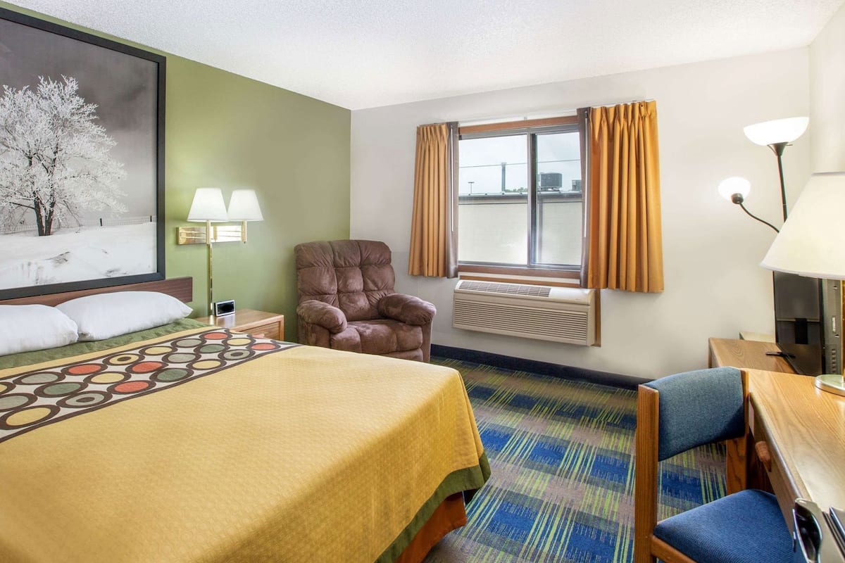 A hotel room with a bed, a chair, a desk, a TV, a window with curtains, an air conditioner, and a painting on the wall.