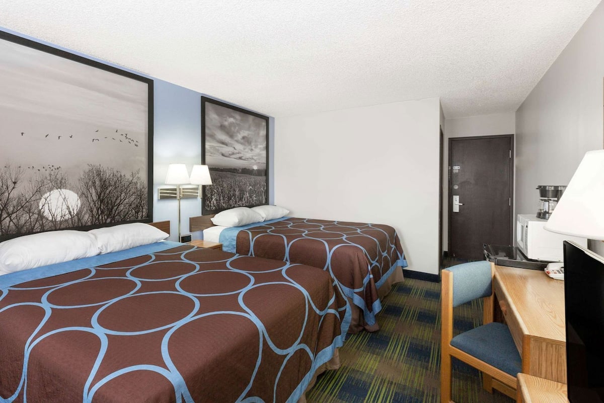 A hotel room with two beds, a desk, a chair, a TV, and artwork on the walls.