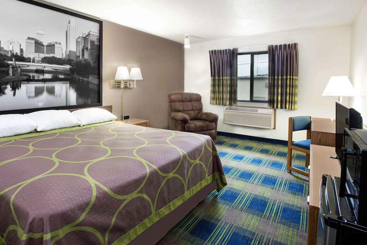 A hotel room with a large bed, a chair, a desk with a computer, a flat-screen TV, and a window with curtains.