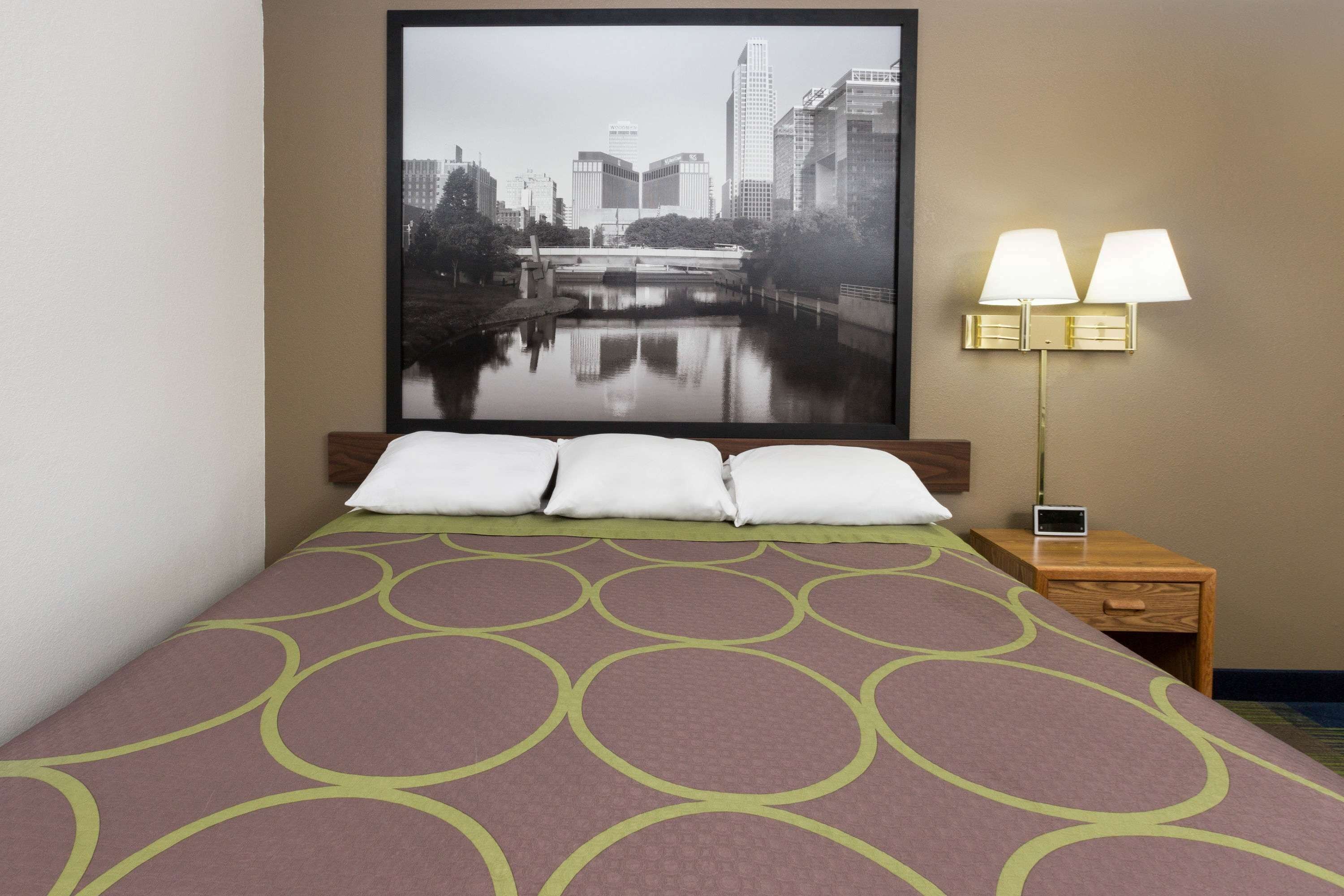 Habitación de hotel con cama, mesita de noche con dos lámparas, cuadro enmarcado con una vista urbana y ventana con vistas al río y edificios.