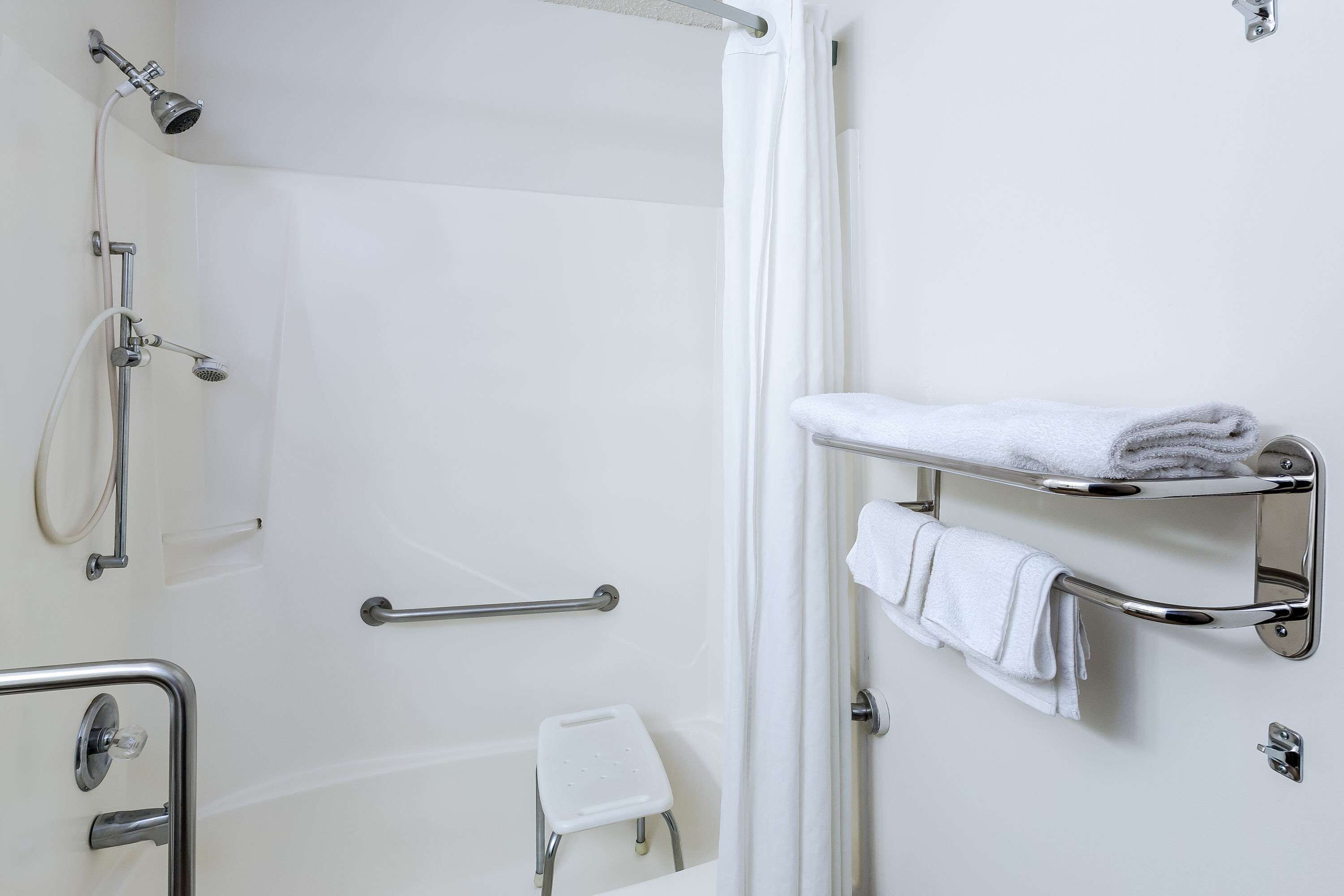 A bathroom with a shower, grab bars, a white towel rack, and a small white stool.