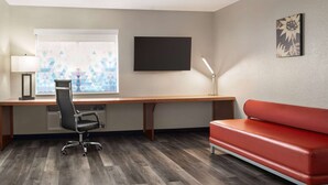 A room with a wooden desk, a black office chair, a red sofa, a TV mounted on the wall, and a window with geometric blinds. - La Quinta Inn & Suites by Wyndham Chicago Tinley Park (Tinley Park)