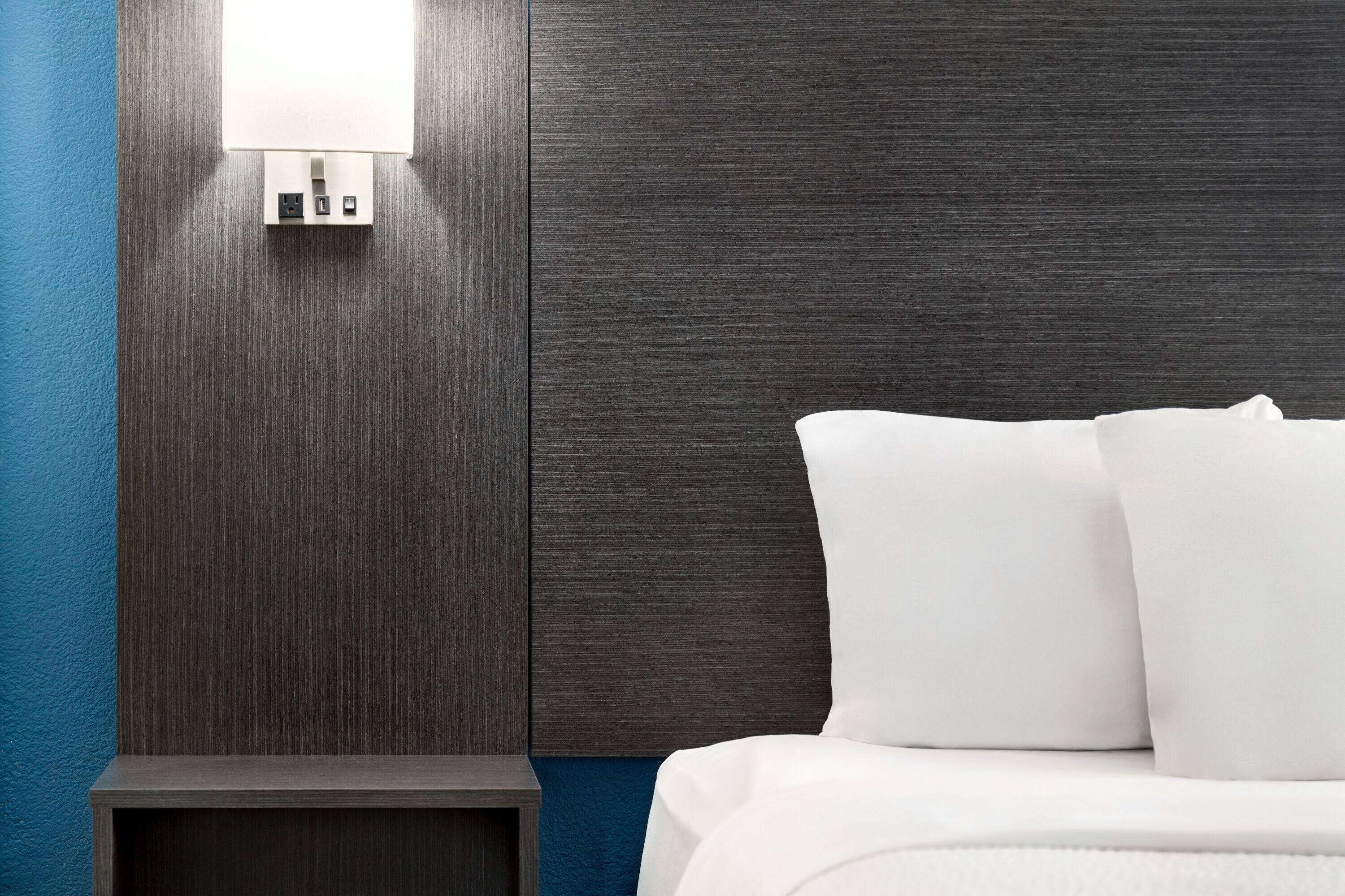 A hotel room with a dark wood headboard, a bedside table, and a white bed with pillows.