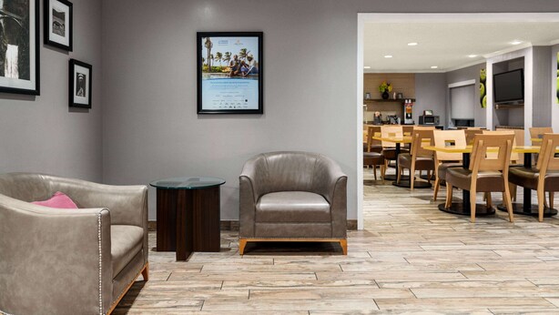 A modern living room with a grey sofa, a leather armchair, and a coffee table. There is a framed certificate on the wall and a dining area with wooden chairs and tables in the background. - La Quinta Inn & Suites by Wyndham Chicago Tinley Park (Tinley Park)